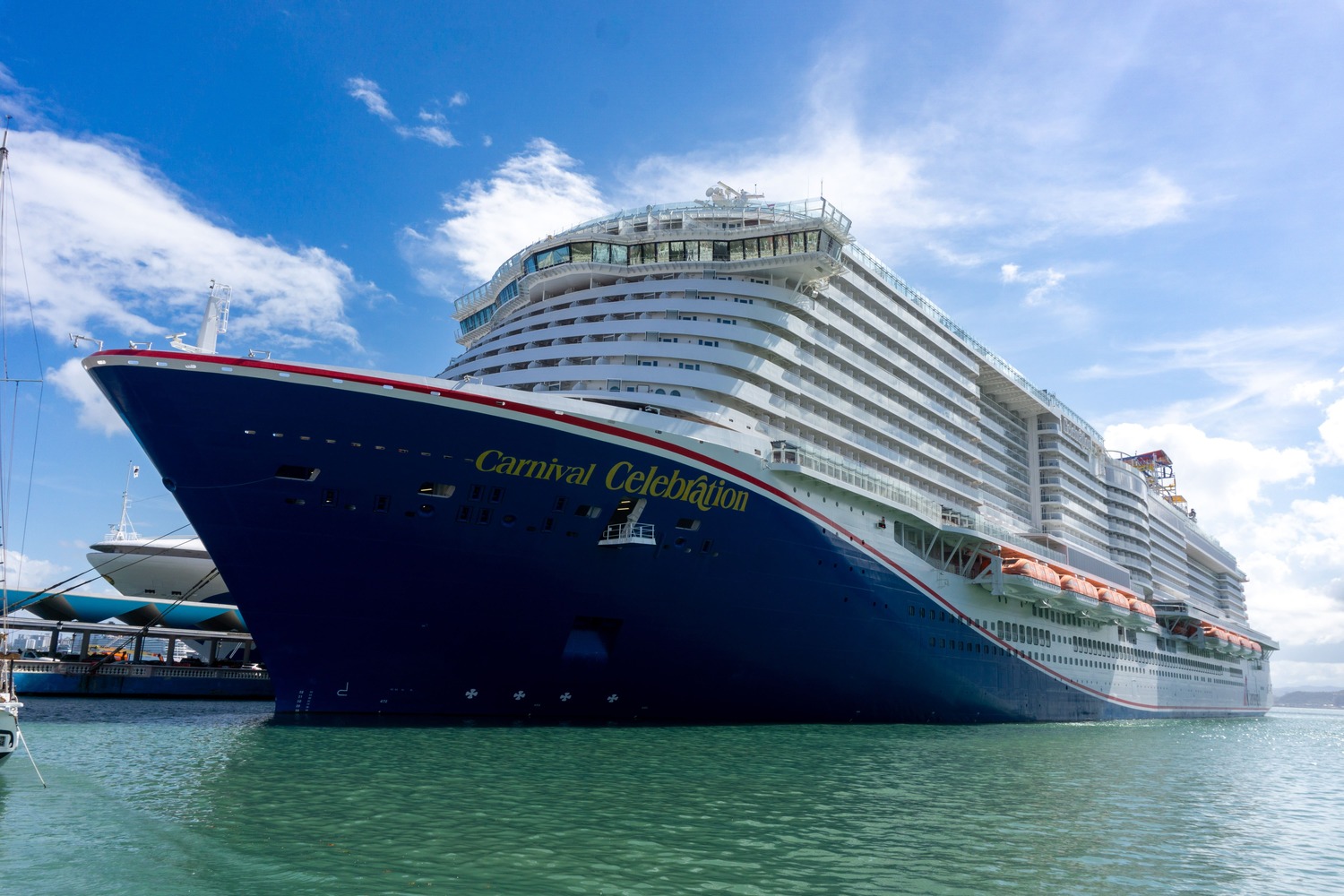 Travel X A large cruise ship named "Carnival Celebration" docked in a harbor on a sunny day, with clear skies and calm waters. Book your trip today to experience the perfect vacation with Travel X. Book Your Trip With Travel X