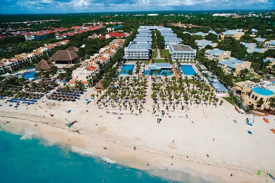 Travel X Aerial view of a beachfront resort with multiple buildings, pools, palm trees, and beach chairs along the sandy shore. The ocean is in the foreground and lush greenery is in the background. Book your trip now for an unforgettable vacation! Book Your Trip With Travel X