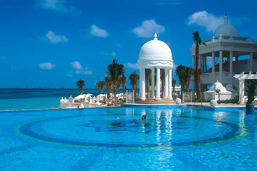 Travel X An outdoor pool with a circular design, surrounded by palm trees and white domed structures, overlooks a calm blue ocean under a clear sky—perfect for your next vacation. Book your trip to this serene resort today! Book Your Trip With Travel X