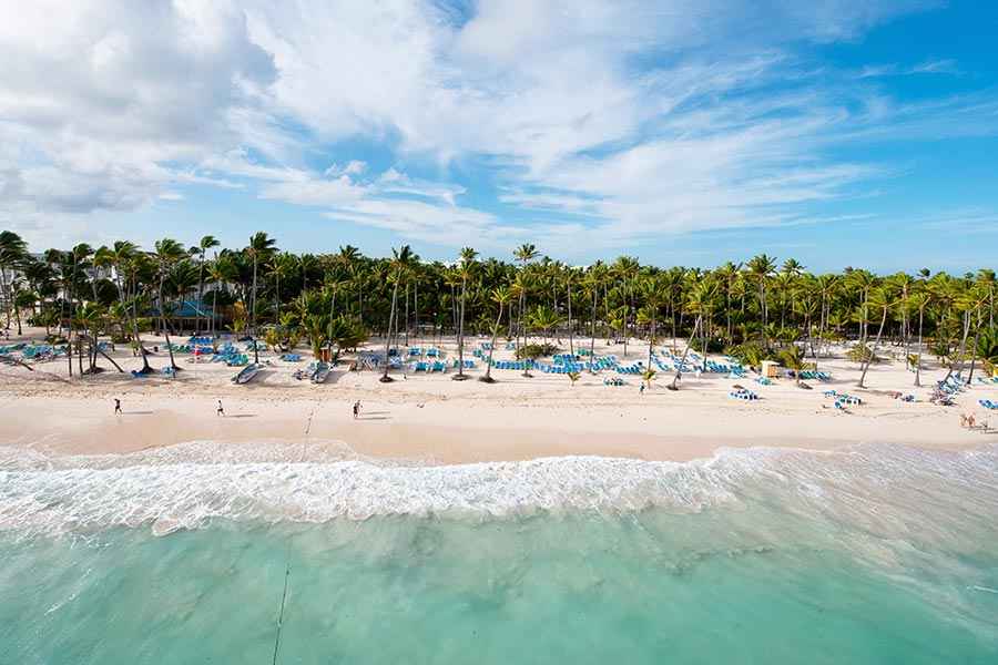 Travel X Aerial view of a sunny beach with turquoise water, white sand, and numerous lounge chairs and palm trees. People are walking along the shore, soaking in the perfect vacation vibe. Book your trip with Travel X today! Book Your Trip With Travel X