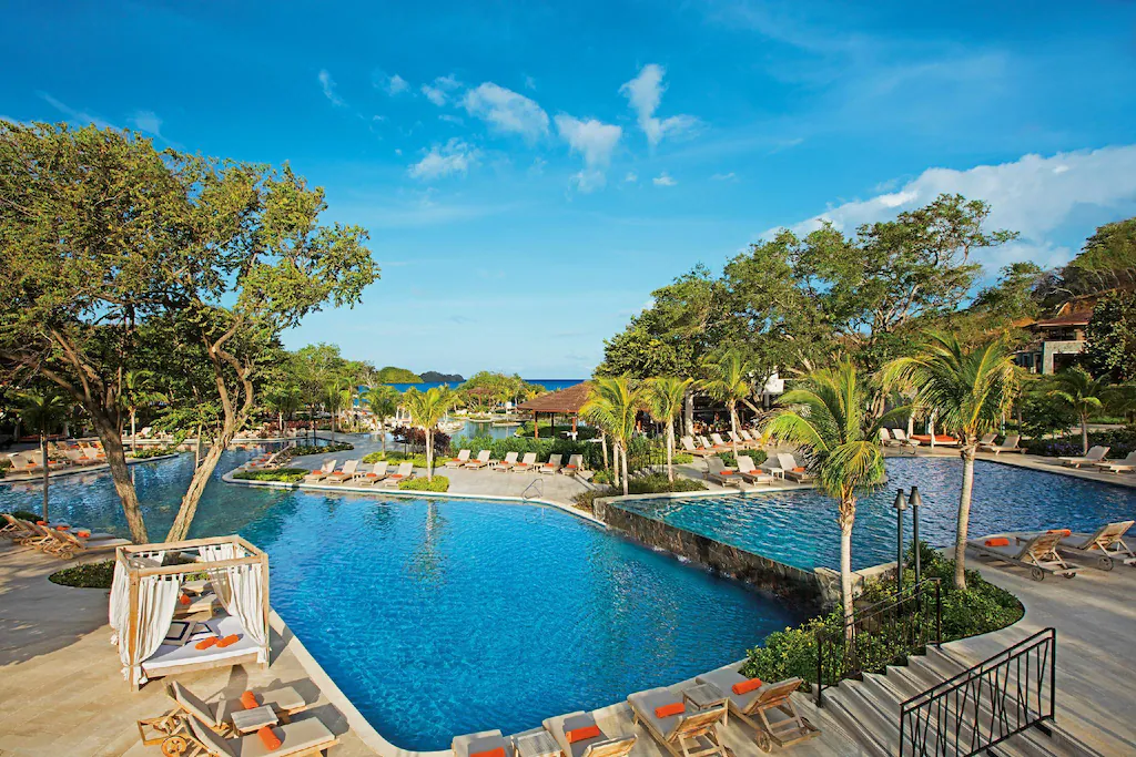 Travel X A resort pool area with lounge chairs, palm trees, and a large swimming pool under a clear blue sky invites you to unwind. Book your trip with Travel X today and transform your vacation dreams into reality. Book Your Trip With Travel X