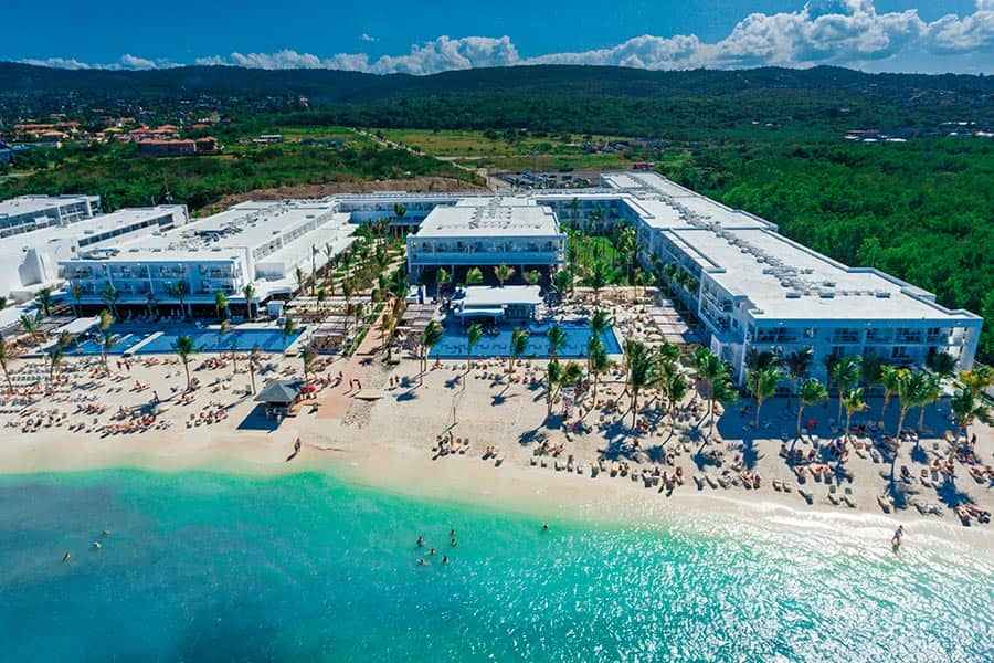 Travel X Aerial view of a large beachfront resort with multiple buildings, swimming pools, palm trees, and guests enjoying the beach and clear turquoise water. Book your trip with Travel X for the ultimate vacation experience! Book Your Trip With Travel X