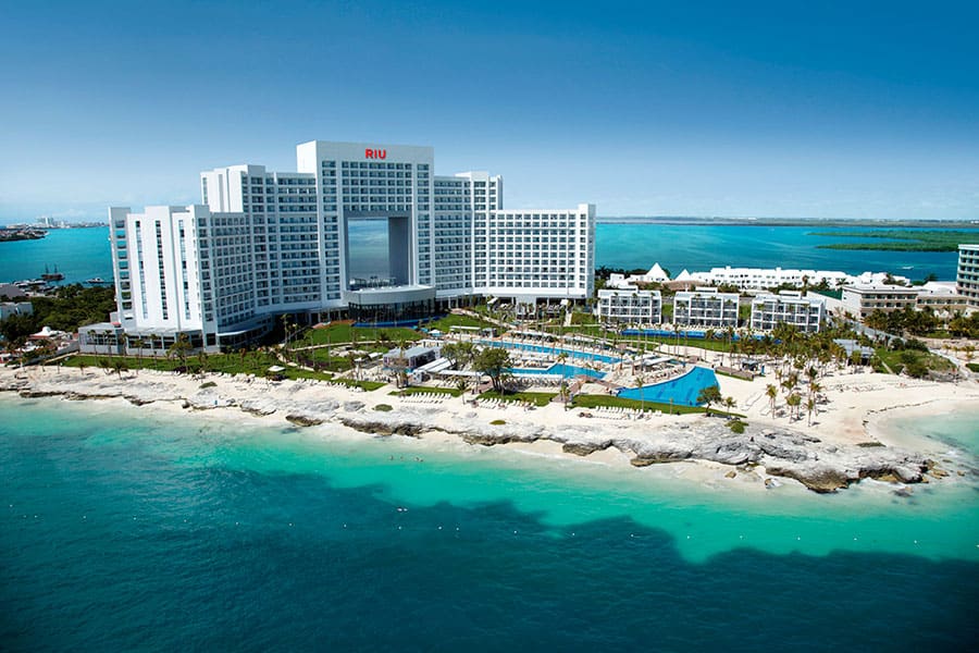 Travel X Aerial view of a large beachfront hotel with multiple swimming pools, surrounded by clear turquoise water and a rocky shoreline. The hotel, perfect for your next Travel X vacation, is situated on a coastal area with a vast ocean in the background. Book your trip now for an unforgettable experience! Book Your Trip With Travel X