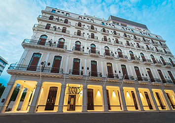 Travel X A multi-story resort with numerous balconies and arched entrances stands against a blue sky background. The building has a symmetrical, formal architectural style and is illuminated from below. Book your trip today to experience this stunning vacation destination! Book Your Trip With Travel X