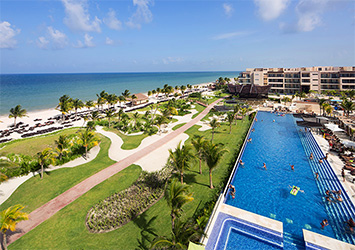 Travel X Aerial view of a beachfront resort featuring a long rectangular pool, lush green lawns, palm trees, and a row of lounge chairs by the ocean under a partially cloudy sky. Perfect for your next Vacation, Book Your Trip with Travel X today! Book Your Trip With Travel X