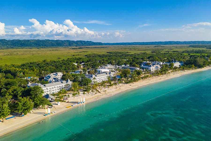 Travel X Aerial view of a coastal vacation resort town with white buildings, sandy beaches, and turquoise waters. Forested hills and clear skies are visible in the background. Book your trip today! Book Your Trip With Travel X