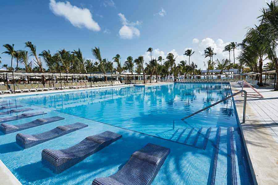 Travel X A large outdoor pool with submerged lounge chairs, surrounded by palm trees under a clear blue sky, epitomizes the perfect resort experience for any vacation. Book Your Trip With Travel X