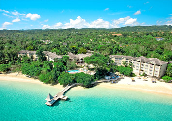 Travel X Aerial view of Travel X beachfront resort surrounded by lush greenery, with clear turquoise water, sandy shore, and a small jetty extending into the sea. Book your trip now to experience this paradise firsthand! Book Your Trip With Travel X