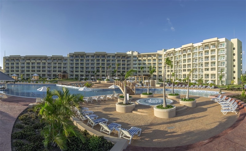Travel X A large resort with multiple floors and numerous balconies, featuring a spacious pool area surrounded by lounge chairs, palm trees, and a bridge over the pool under a clear blue sky. Ready for the perfect vacation? Book your trip today! Book Your Trip With Travel X