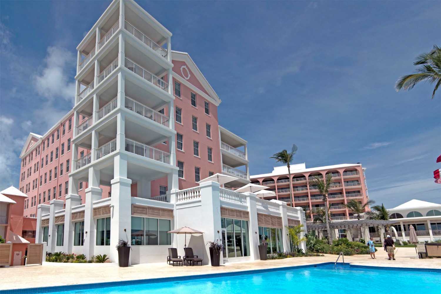 Travel X A multi-story pink resort with balconies overlooks a blue swimming pool. There are lounge chairs and umbrellas around the pool area under a clear sky. Palm trees are seen in the background, offering a perfect vacation setting. Book your trip now to bask in this tropical paradise! Book Your Trip With Travel X