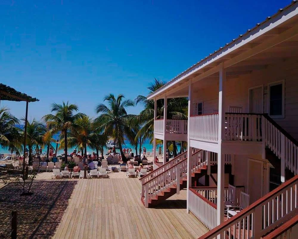 Travel X A beachfront resort featuring wooden buildings, multiple palm trees, and lounge chairs facing the ocean under a clear blue sky. Book your trip with Travel X today for the ultimate coastal getaway! Book Your Trip With Travel X