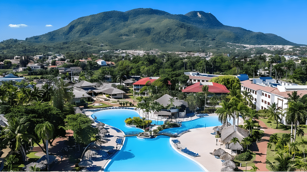 Travel X Aerial view of a Travel X resort featuring multiple blue pools, surrounded by palm trees and buildings, with a backdrop of green mountains under a clear blue sky. Book Your Trip now for an unforgettable getaway! Book Your Trip With Travel X