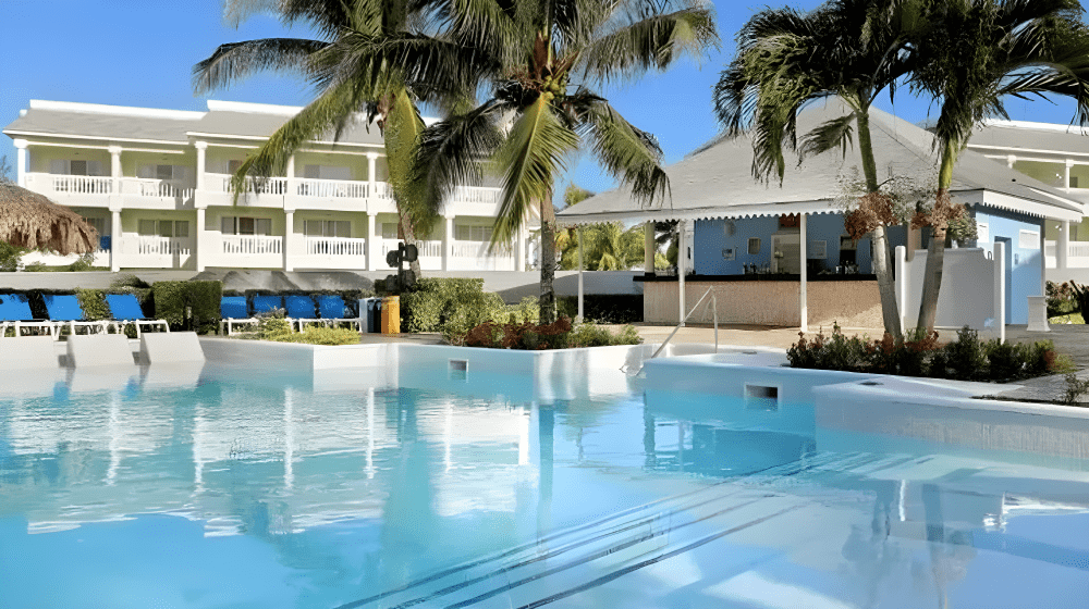 Travel X A clear pool surrounded by palm trees, lounge chairs, and a two-story white building with balconies sets the scene for your dream vacation. A small bar shaded by a pavilion is on the right. Book your trip to this serene resort today! Book Your Trip With Travel X