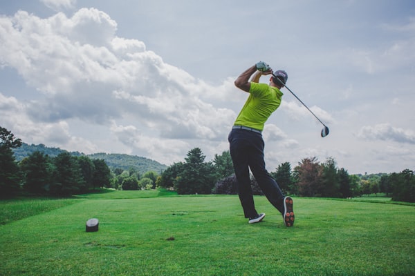 Travel X A golfer in a yellow shirt swings a club on a grassy golf course with trees and hills in the background under a partly cloudy sky, painting the perfect backdrop for your next Vacation. Book Your Trip with Travel X and experience this scenic beauty firsthand. Book Your Trip With Travel X