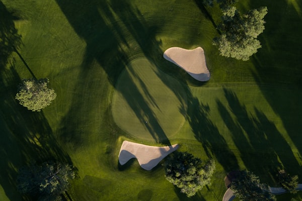 Travel X Aerial view of a golf course green with two sand bunkers, surrounded by trees casting long shadows—an inviting scene that makes you want to book your trip for a relaxing vacation at this stunning resort. Book Your Trip With Travel X