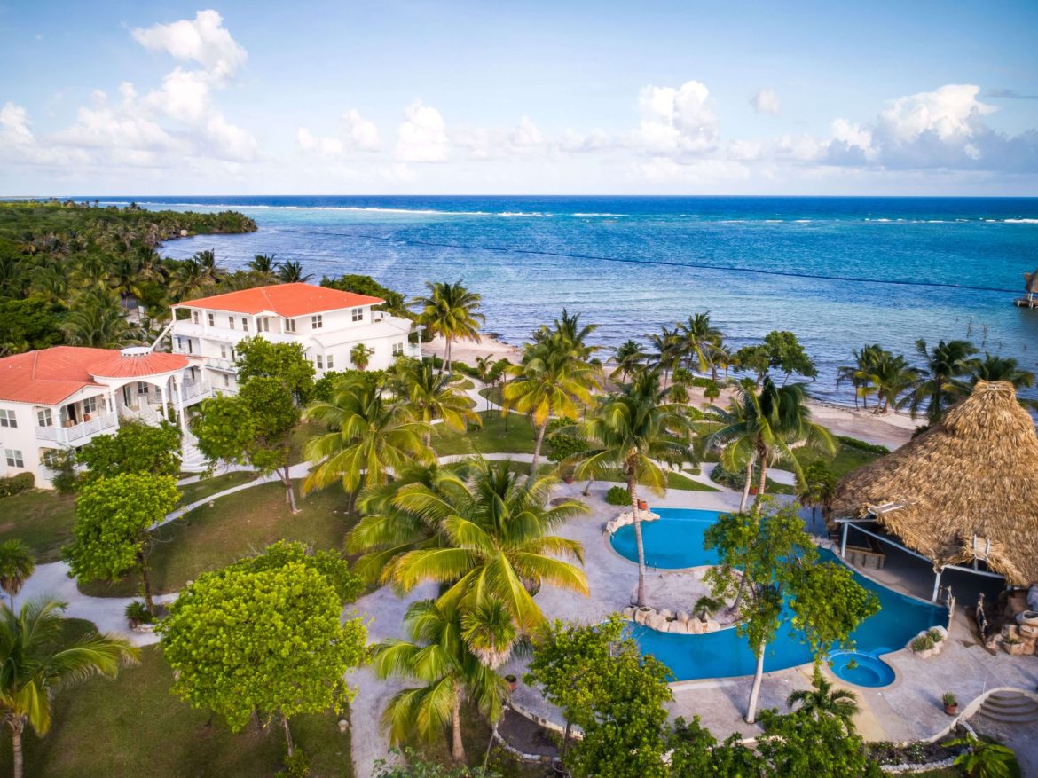 Travel X Aerial view of a beachfront resort with white buildings, red roofs, palm trees, a blue swimming pool, and a thatched structure against the ocean backdrop—perfect for your next vacation. Book Your Trip with Travel X today! Book Your Trip With Travel X