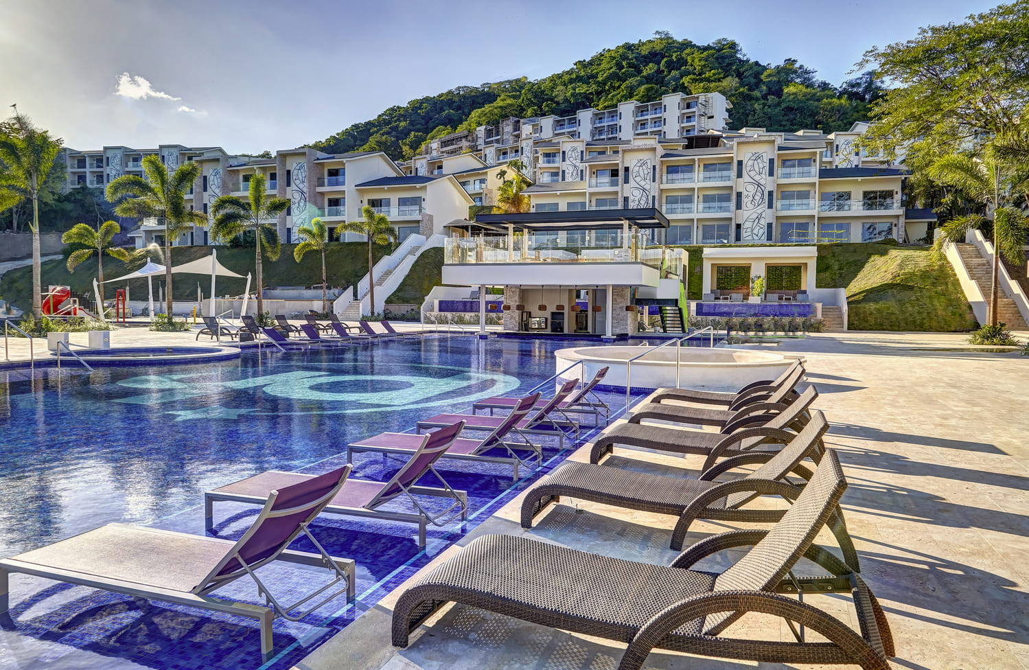 Travel X Experience the ultimate Resort getaway with a luxurious pool area, lounge chairs, shaded cabanas, and modern buildings framed by lush hills under a clear sky. Book your trip today with Travel X and make unforgettable memories! Book Your Trip With Travel X