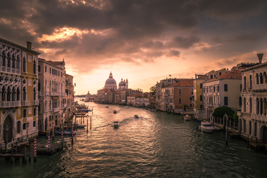 Travel X A canal in Venice at sunset with boats moving along the water, bordered by historic buildings and a cathedral dome in the background under a partly cloudy sky—a perfect setting for your next vacation. Book your trip to this enchanting resort city today! Book Your Trip With Travel X