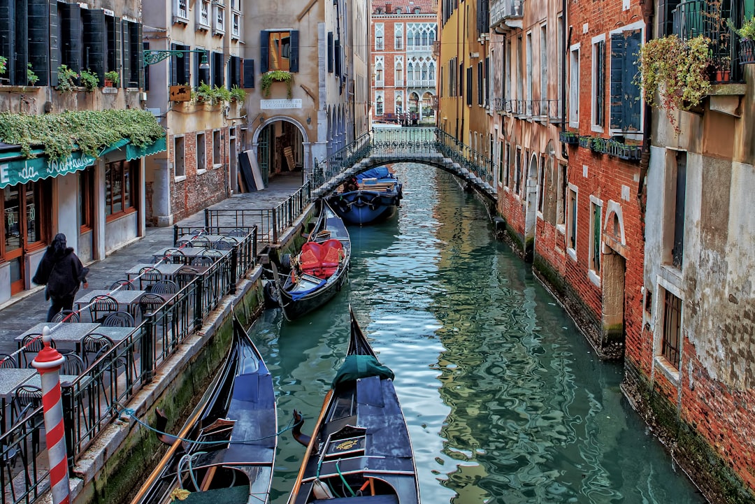 Travel X A narrow canal in Venice, with gondolas moored along the sides. A small bridge arches over the canal, and buildings with balconies line the waterway as a person walks on the sidewalk. It's a picture-perfect vacation scene—Book Your Trip now! Book Your Trip With Travel X