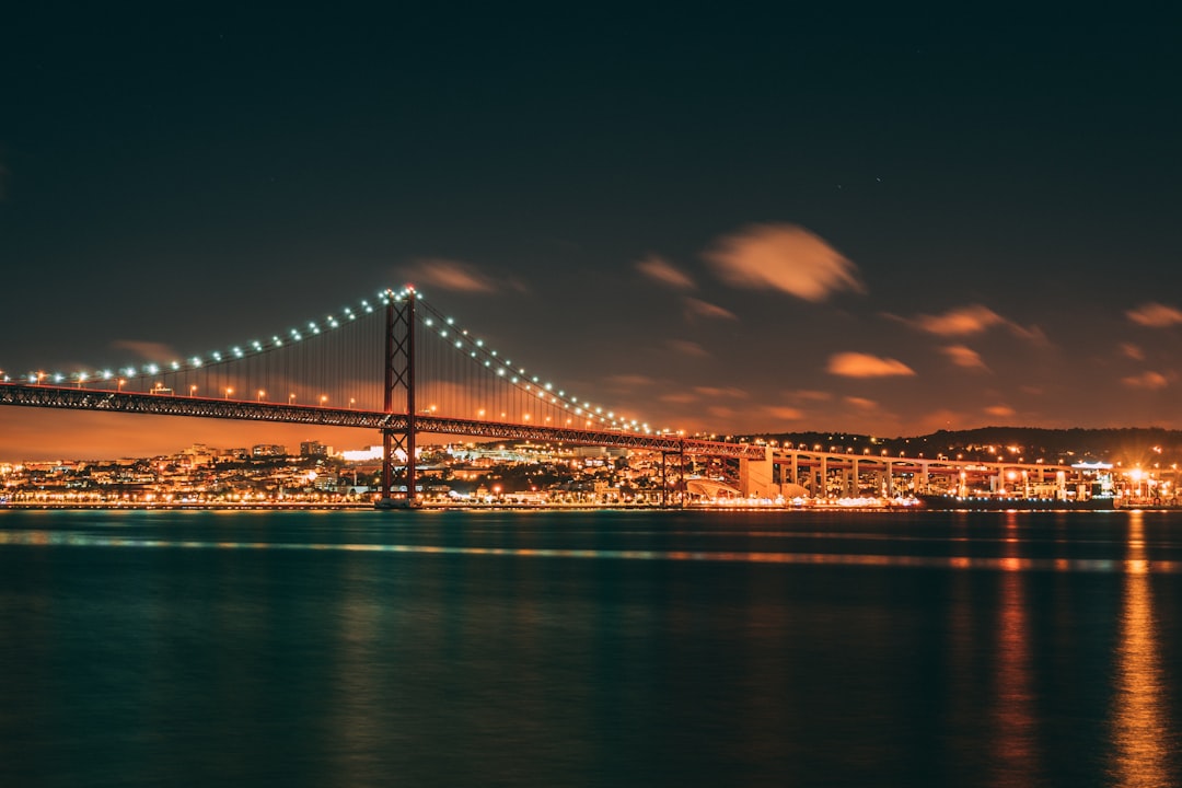 Travel X A suspension bridge is illuminated at night over calm waters with the city lights reflecting on the water's surface under a clear sky, showcasing a perfect evening setting for your next vacation. Book your trip now to experience this stunning resort-like scenery firsthand. Book Your Trip With Travel X