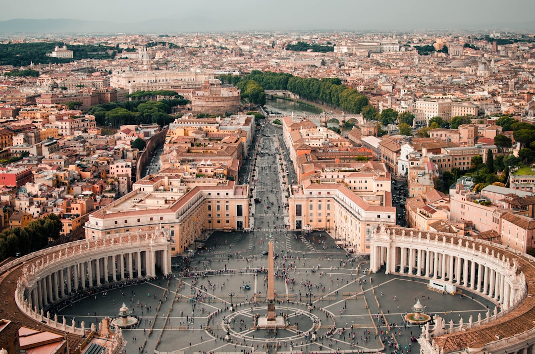 Travel X Aerial view of St. Peter's Square in Vatican City, showcasing its symmetrical colonnades, central obelisk, and surrounding cityscape. The Tiber River is visible in the background—a perfect snapshot for your travel album during a dream vacation. Book your trip with Travel X today! Book Your Trip With Travel X