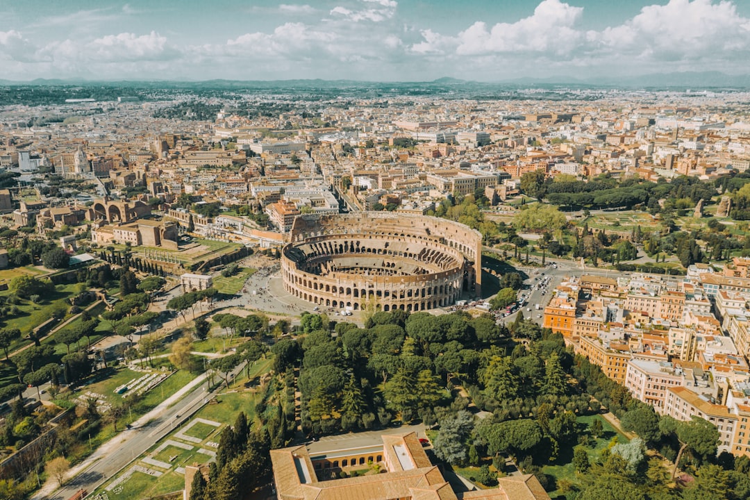 Travel X Aerial view of the Colosseum in Rome, surrounded by city buildings and green spaces under a sunny sky with clouds, creates a picture-perfect scene for any Travel X vacation itinerary. Book Your Trip With Travel X