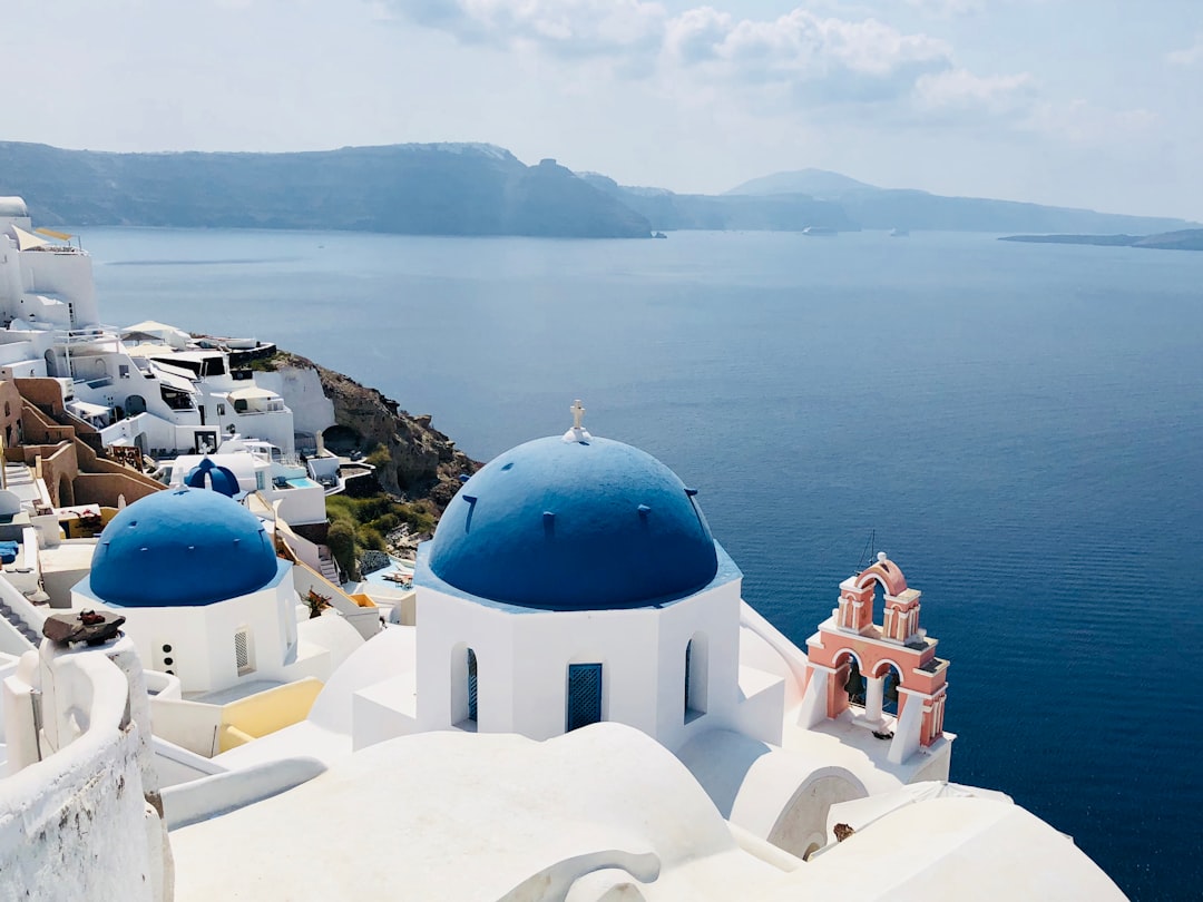 Travel X View of white buildings with blue domes and a pink bell tower overlooking the sea in Santorini, Greece, with cliffs in the background. This picture-perfect resort is the ideal backdrop for your dream vacation. Book your trip today to experience its breathtaking beauty firsthand! Book Your Trip With Travel X