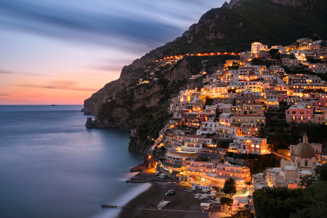 Travel X A coastal town built on a hillside is illuminated at dusk, with the sea to the left and mountains in the background—an ideal resort destination for your next vacation. Book Your Trip With Travel X