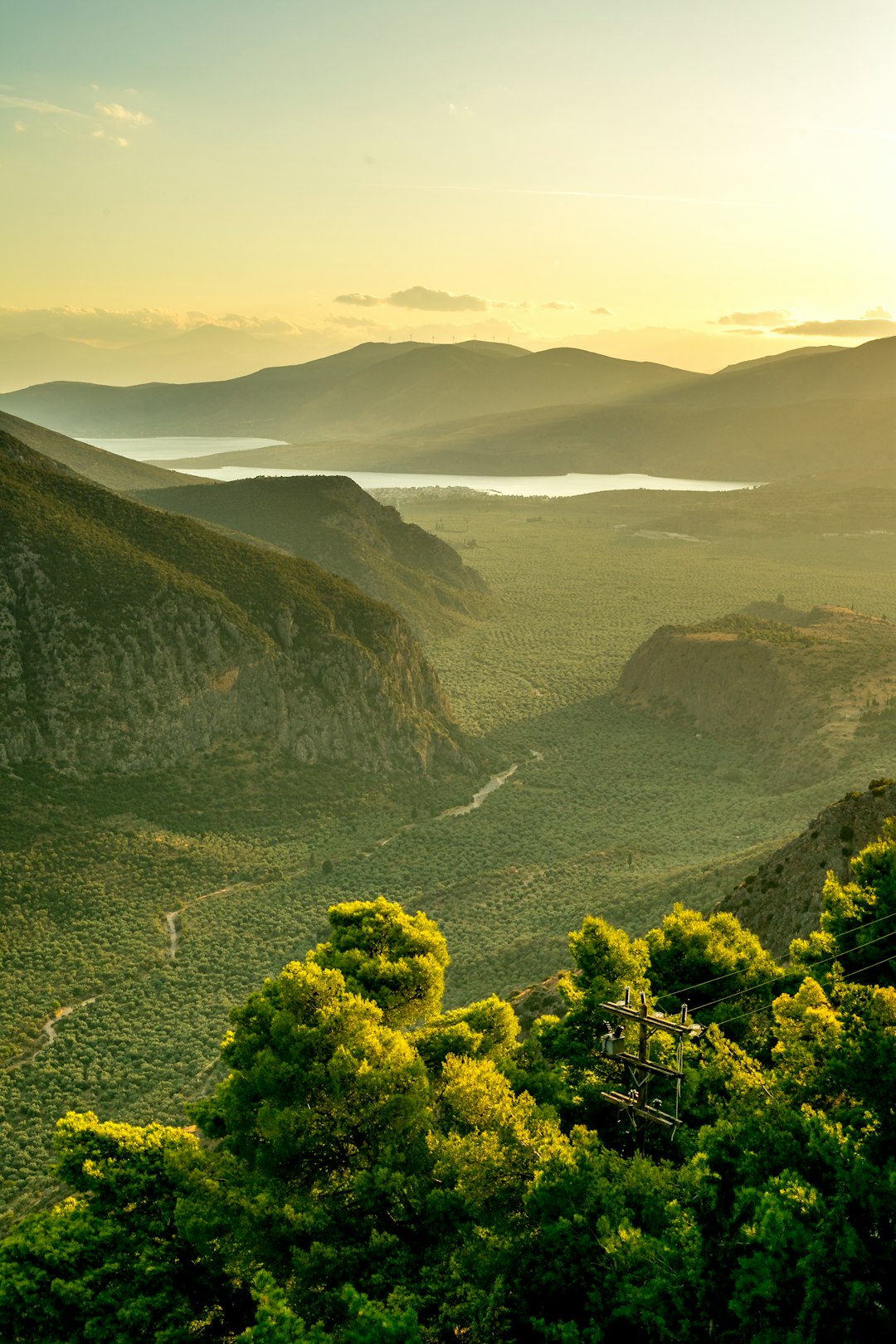 Travel X Aerial view of a lush green valley flanked by mountains, with a river running through it and a hazy backdrop of hills and a lake under a golden sunset—an ideal vacation spot that rivals any resort. Book Your Trip With Travel X