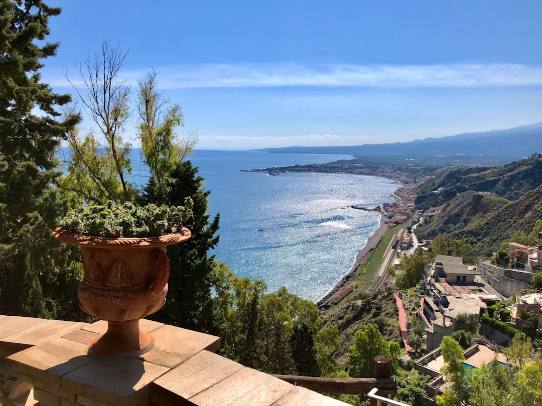 Travel X A terracotta planter with greenery overlooks a coastal town, with the expansive sea, shoreline, and hills visible under a clear blue sky—a perfect scene to inspire you to book your trip for an unforgettable vacation. Book Your Trip With Travel X