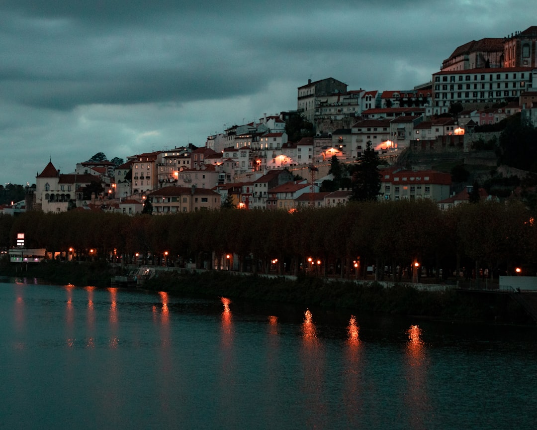 Travel X Hilltop town with lit buildings at dusk, reflecting on a calm river under a cloudy sky. Experience this enchanting destination with Travel X. Book your trip to this serene resort and capture the magic today. Book Your Trip With Travel X