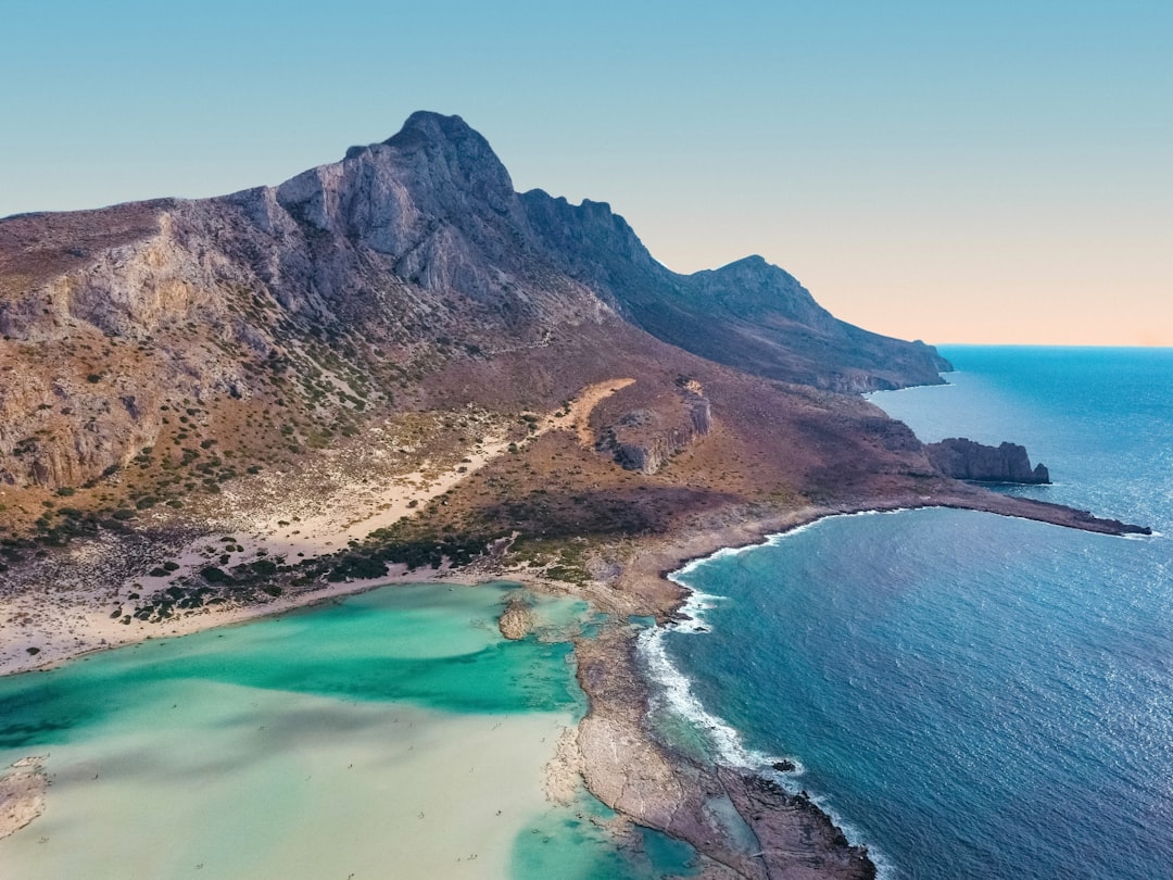 Travel X Aerial view of a coastal landscape featuring a rugged mountain range and a coastline with turquoise and deep blue waters—a stunning natural retreat perfect for your next Travel X vacation. Book Your Trip With Travel X