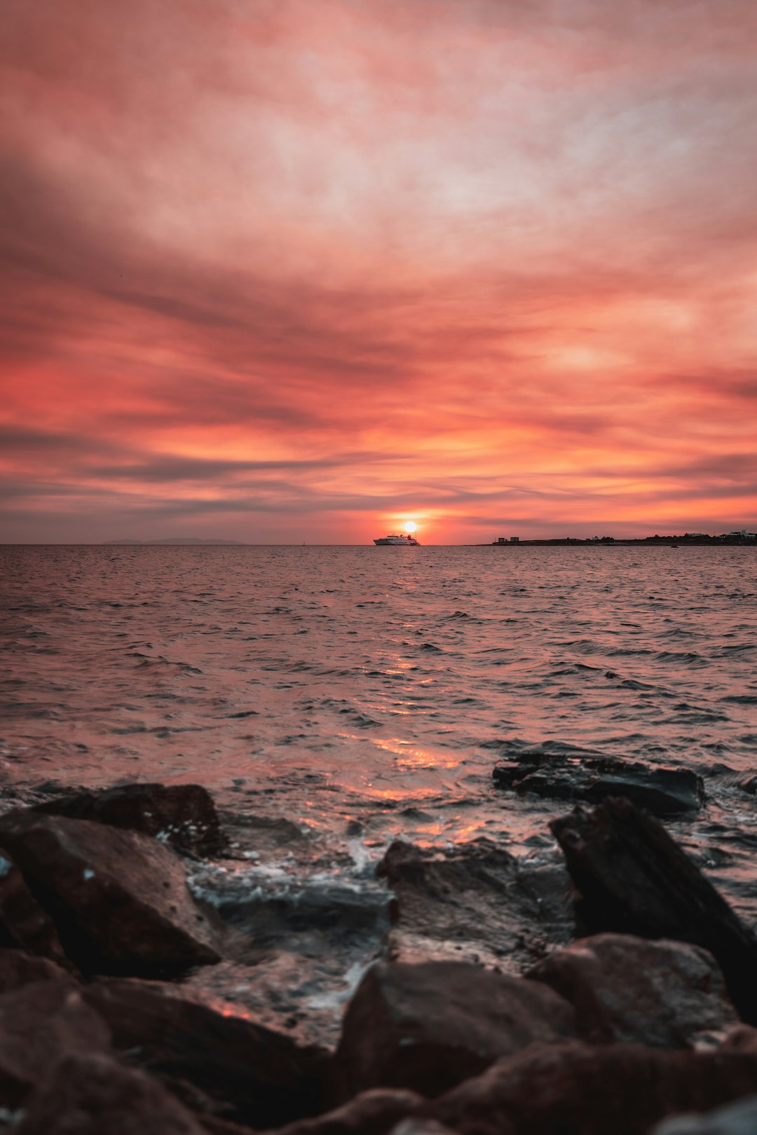 Travel X A sunset over a body of water with a ship on the horizon. The sky is filled with vibrant red and orange hues, and rocks are visible in the foreground by the shore, creating a perfect Travel X moment that makes you dream of your next vacation. Book Your Trip With Travel X