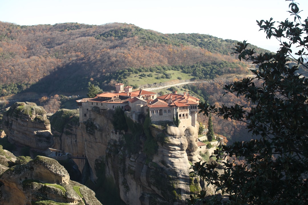 Travel X A large monastery complex sits atop a rocky cliff surrounded by a mountainous landscape with trees showing autumn colors. A distant road is visible behind the monastery, inviting travelers to dream of their next getaway. Book your trip today with Travel X for an unforgettable vacation. Book Your Trip With Travel X