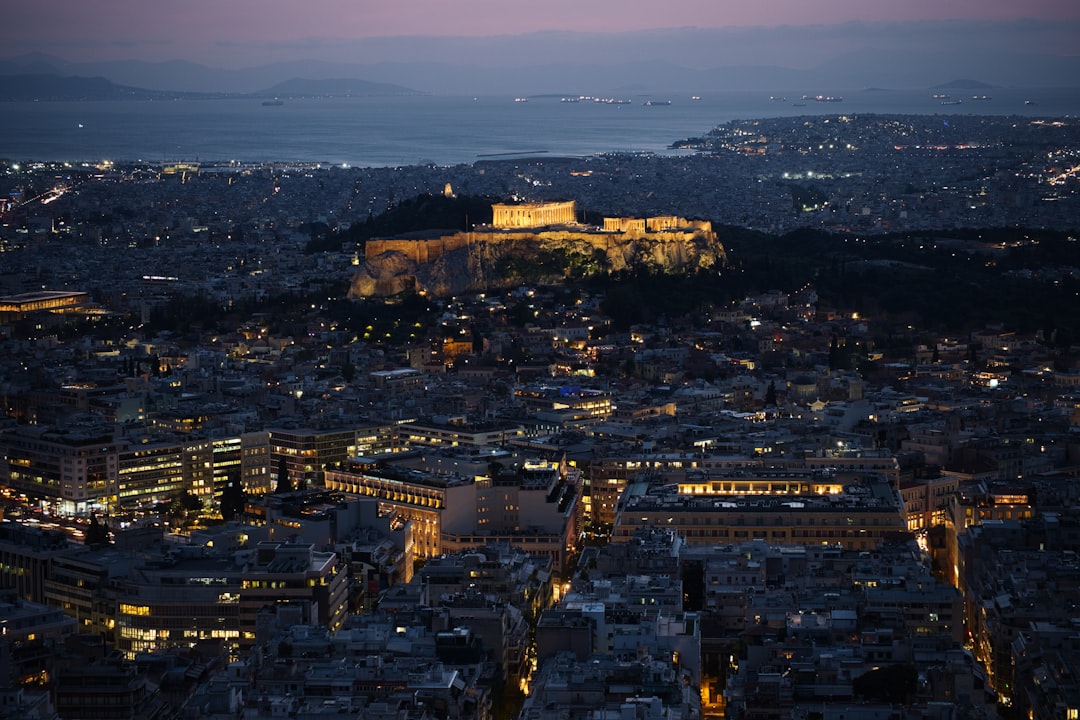 Travel X Aerial view of Athens at dusk, with the illuminated Acropolis prominently visible in the center, and densely packed city buildings surrounding it. Travel X offers vacation packages that showcase this stunning scene, perfect for those seeking a blend of history and modernity. Book Your Trip With Travel X