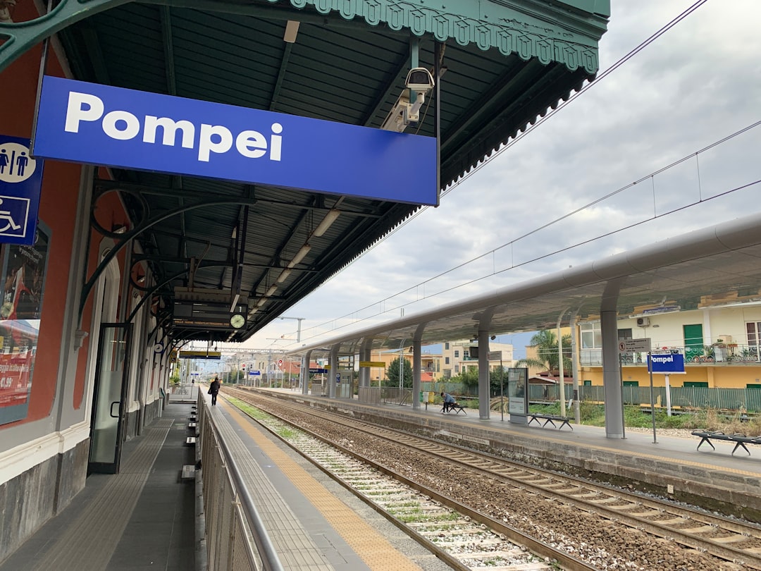 Travel X Outdoor railway station platform in Pompei, Italy, with tracks and a blue station sign reading "Pompei." The platform is empty with buildings visible in the distance. Plan your next getaway with Travel X and book your trip to this charming destination today! Book Your Trip With Travel X