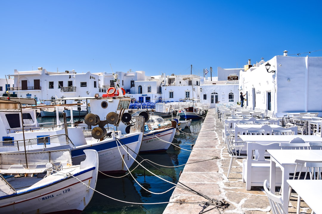 Travel X A coastal scene featuring several boats docked at a marina with a stone walkway lined with white tables and chairs next to whitewashed buildings under a clear blue sky, perfect for a Travel X vacation. Book Your Trip With Travel X