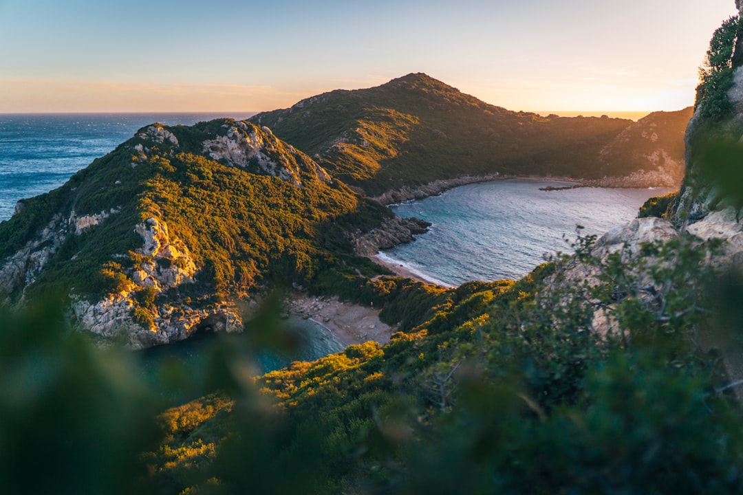 Travel X Aerial view of a coastal landscape with green hills surrounding a crescent-shaped bay at sunset, perfect for your next vacation. Book Your Trip With Travel X