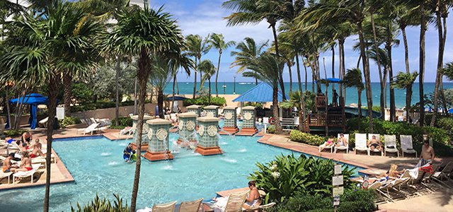 Travel X The resort's outdoor pool, surrounded by palm trees and lounge chairs, offers a picturesque vacation setting with people relaxing and a stunning view of the ocean in the background. Book Your Trip With Travel X