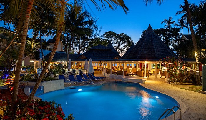 Travel X Vacation under the stars with a nighttime view of a resort featuring a lit swimming pool, surrounded by lounge chairs and palm trees. The building with outdoor seating in the background completes this serene escape. Book your trip today for the ultimate relaxation experience. Book Your Trip With Travel X
