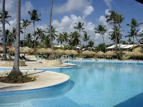 Travel X A large, clear blue swimming pool with palm trees and thatched umbrellas surrounding it under a partly cloudy sky creates the perfect vacation vibe at this luxurious resort. Book Your Trip With Travel X