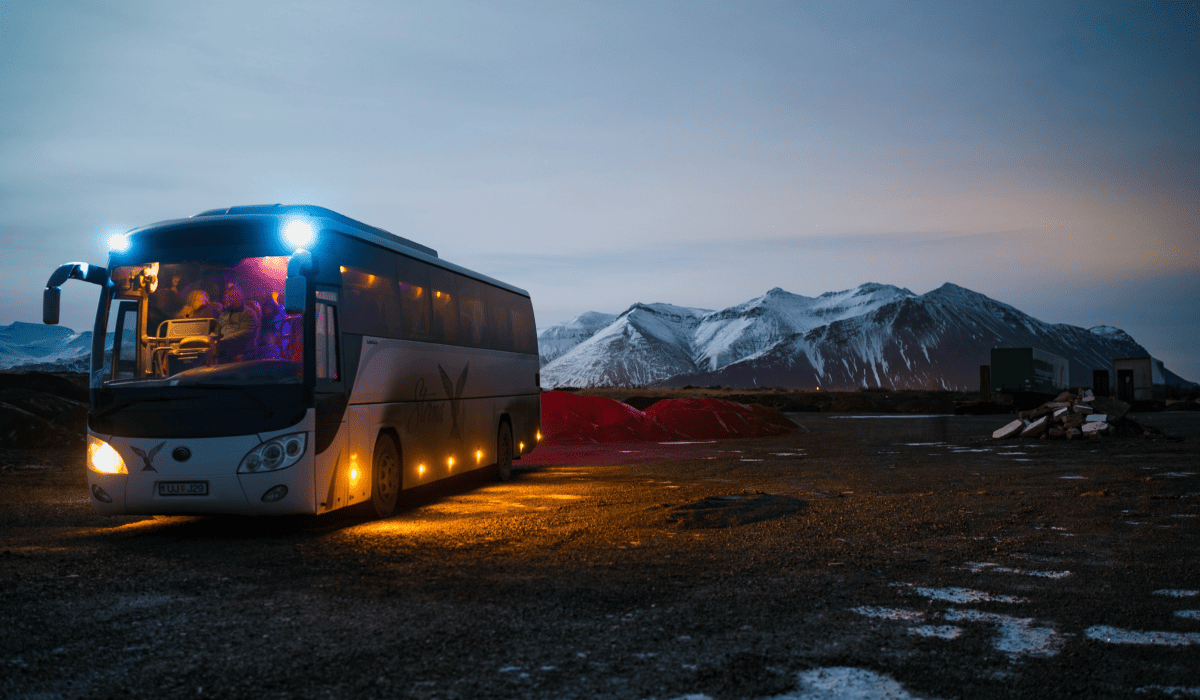 Travel X A Travel X tour bus with lights on is parked on a gravel area at dusk, with a snow-capped mountain range in the background. Book your trip today to experience this breathtaking view! Book Your Trip With Travel X