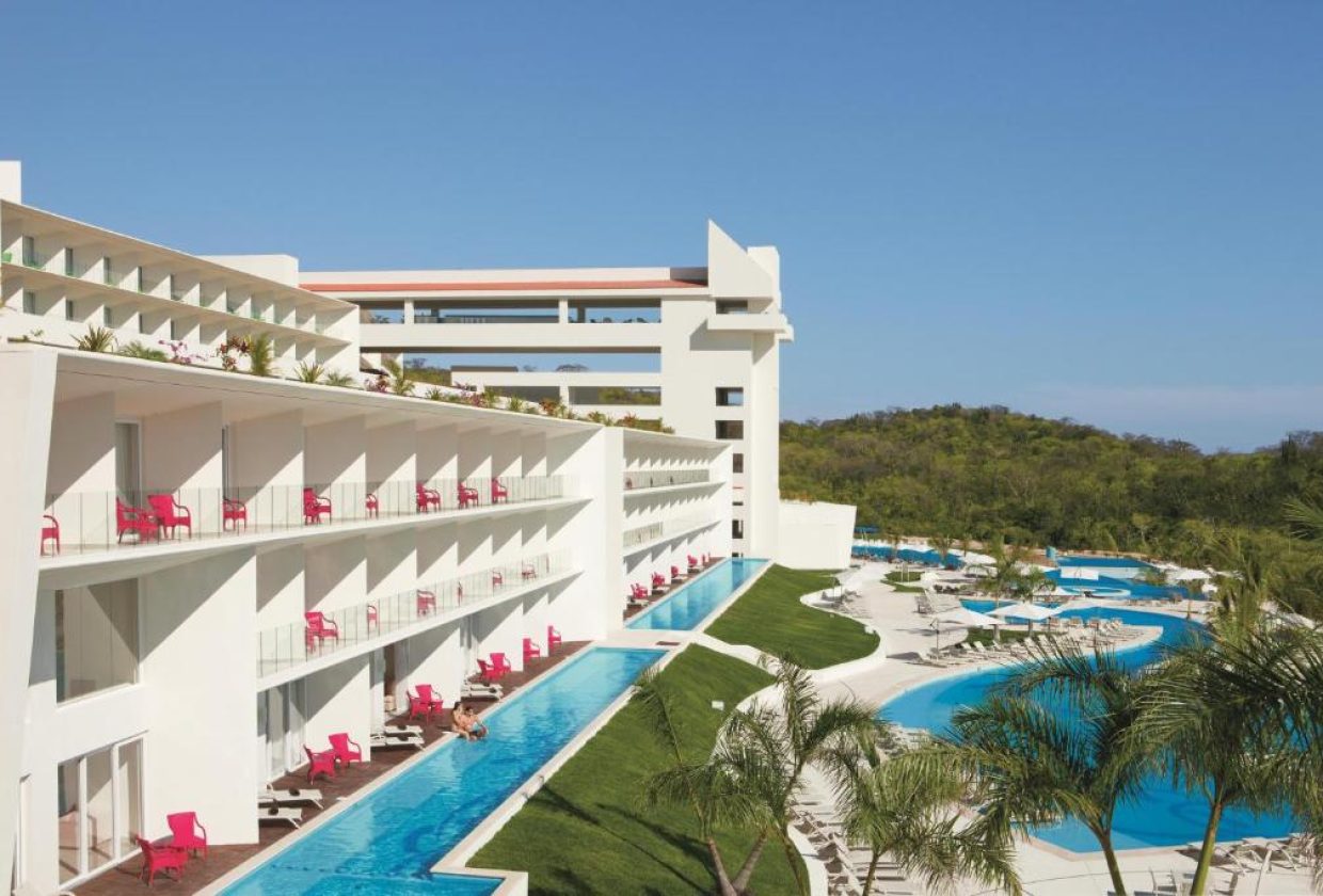 Travel X Modern white building with red chairs on balconies beside a long, narrow pool. The structure overlooks a larger curved pool with white lounge chairs and palm trees, perfect for a Travel X vacation. Blue sky and greenery in the background enhance this stunning resort scene. Book Your Trip With Travel X