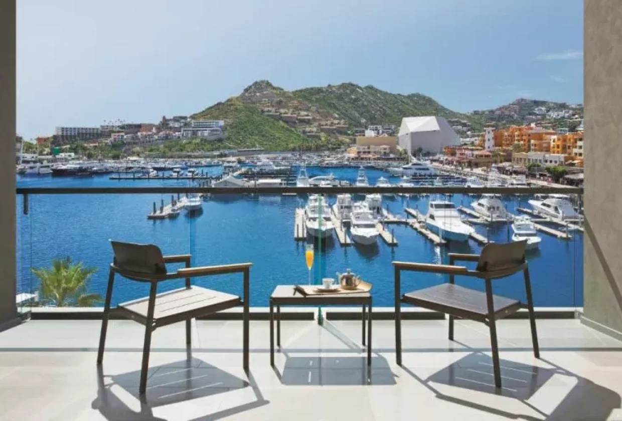 Travel X Two chairs and a table with a tea set on a balcony at the resort, overlooking a marina filled with boats and yachts, surrounded by mountains and buildings. Book Your Trip With Travel X