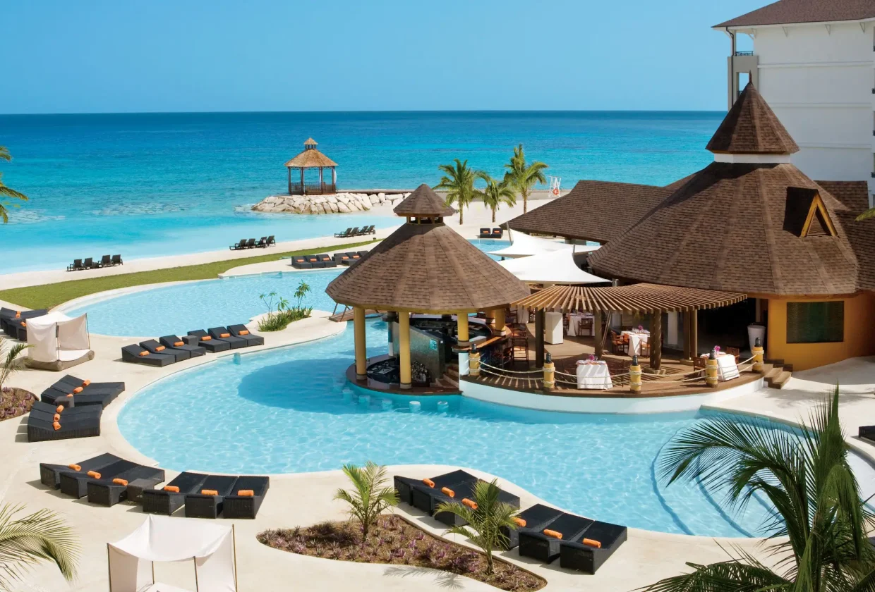 Travel X A tropical resort with a large pool, poolside cabanas, a swim-up bar, and a gazebo near the ocean under a clear blue sky. Book your trip with Travel X for the ultimate vacation. Book Your Trip With Travel X