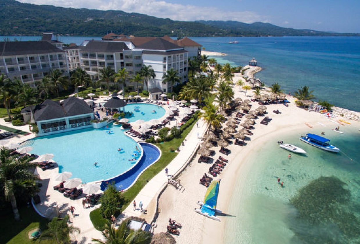 Travel X Aerial view of a Travel X beachfront resort featuring multiple pools, sun loungers, palm trees, and a docked boat in a stunning tropical setting. Perfect for your dream vacation. Book Your Trip With Travel X