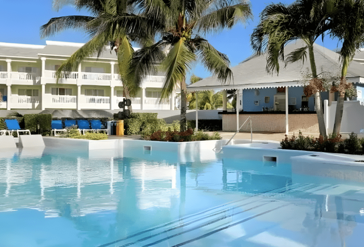 Travel X A clear pool surrounded by palm trees, lounge chairs, and a two-story white building with balconies sets the scene for your dream vacation. A small bar shaded by a pavilion is on the right. Book your trip to this serene resort today! Book Your Trip With Travel X